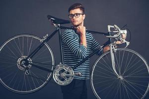 in seinem eigenen Stil. Selbstbewusster junger gutaussehender Mann mit Brille, der sein Fahrrad im Retro-Stil auf der Schulter trägt und wegschaut, während er vor grauem Hintergrund steht foto