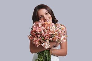 er liebt mich definitiv. Attraktive junge Frau mit Blumenstrauß, die Kamera anschaut und lächelt, während sie vor grauem Hintergrund steht foto