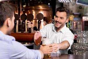 Hier ist dein Bier. fröhlicher junger barkeeper, der dem kunden einen becher mit bier gibt foto