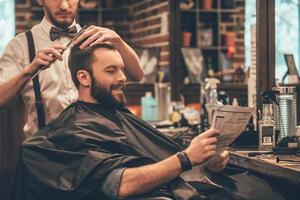 tolle Zeit beim Friseur. Fröhlicher junger bärtiger Mann, der sich vom Friseur die Haare schneiden lässt und Zeitung liest, während er im Friseursalon auf dem Stuhl sitzt foto