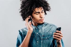 schöner Takt. junger afrikanischer mann, der kopfhörer trägt und sein smartphone hält, während er die augen vor grauem hintergrund geschlossen hält foto