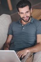 gerne zu Hause arbeiten. Blick aus der Vogelperspektive auf einen gutaussehenden jungen Mann, der seinen Laptop benutzt und mit einem Lächeln in die Kamera schaut, während er zu Hause auf der Couch sitzt
