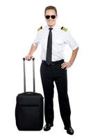 neuer tag und neues ziel. Selbstbewusster männlicher Pilot in Uniform, der die Hand auf seinen Koffer lehnt und lächelt, während er isoliert auf weißem Hintergrund ist foto