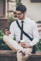 immer in Kontakt. Selbstbewusster junger Mann mit Brille, der eine Kaffeetasse hält und sein Smartphone benutzt, während er im Freien sitzt foto