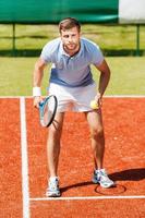 selbstbewusster Tennisspieler. hübscher junger mann im poloshirt, der tennisschläger hält und konzentriert aussieht, während er auf dem tennisplatz steht foto