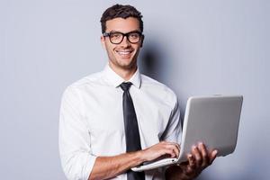 zuversichtlich es Experte. hübscher junger Mann in Hemd und Krawatte, der am Laptop arbeitet und im Stehen vor grauem Hintergrund lächelt foto