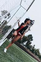 schöner Spieler. volle Länge der attraktiven jungen Frau im roten Bikini, der nahe Torpfosten auf dem Fußballfeld aufwirft foto
