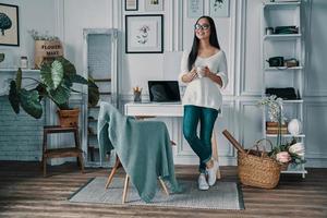 frischen Kaffee genießen. in voller Länge von einer schönen jungen Frau, die wegschaut und lächelt, während sie im Home Office steht foto