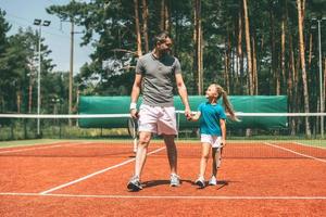 Volle Länge des kleinen blonden Haarmädchens in Sportkleidung, das Tennisschläger trägt und ihren Vater anschaut, der neben ihr am Tennisplatz spazieren geht foto