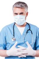 Vorbereitung auf die Operation. reifer Arzt mit grauem Haar in OP-Maske und Handschuhen, der in die Kamera blickt, während er isoliert auf Weiß steht foto