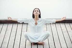 Ruhe und Ausgeglichenheit finden. Schöne junge Frau in weißer Kleidung, die im Lotussitz sitzt und die Augen geschlossen hält, während sie im Freien meditiert foto