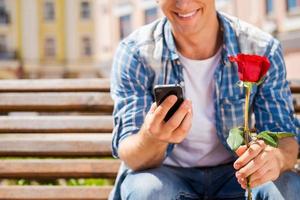 Auf sie warten. Nahaufnahme eines glücklichen jungen Mannes, der eine einzelne Rose und ein Handy hält, während er auf der Bank sitzt foto