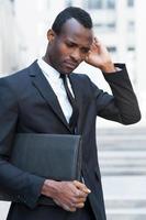 in Gedanken verloren. nachdenklicher junger afrikanischer mann in formalwear, der die hände gefaltet hält und wegschaut, während er im freien sitzt foto