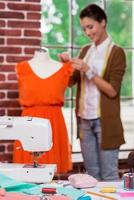 Spaß an kreativer Arbeit. Schöne junge Modedesignerin, die das Kleid auf der Schaufensterpuppe anpasst und mit der Nähmaschine im Vordergrund lächelt foto