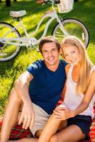 Paar im Park entspannen. glückliches junges Liebespaar, das sich gemeinsam im Park entspannt, während es auf der Picknickdecke und mit dem Fahrrad im Hintergrund sitzt foto