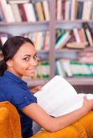 schöner Bücherwurm. Blick von oben auf die schöne afrikanische Studentin, die ein Buch hält und lächelt, während sie auf dem Stuhl in der Bibliothek sitzt foto