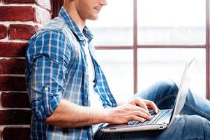 Arbeiten am Laptop. Seitenansicht eines jungen Mannes, der am Laptop arbeitet, während er auf der Fensterbank sitzt foto