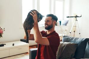 Verspielter junger Mann in Freizeitkleidung, der sich mit Hauskatze verbindet und lächelt, während er Zeit im Haus verbringt foto