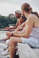 freundliches Gespräch. Gruppe glücklicher junger Menschen in Freizeitkleidung, die lächeln und reden, während sie auf dem Pier sitzen foto