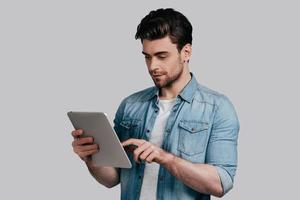 mit modernen Technologien. Gut aussehender junger Mann im Blue Jeans-Hemd, der an einem digitalen Tablet arbeitet, während er vor grauem Hintergrund steht foto