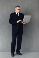 Technologien erleichtern das Geschäft. in voller Länge vom reifen Geschäftsmann, der seinen Laptop verwendet, während er gegen grauen Hintergrund steht foto