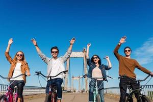 Fahrspaß genießen. Tiefwinkelansicht von vier jungen, fröhlichen Menschen, die ihre Fahrräder fahren und die Arme hochhalten foto