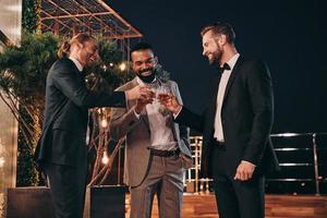 Drei gutaussehende Männer in Anzügen trinken Whiskey und kommunizieren, während sie Zeit auf der Party verbringen foto