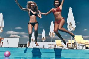 verrückte Mädchen. Zwei attraktive junge Frauen im Bikini halten sich an den Händen und halten den Mund offen, während sie im Freien in den Pool springen foto