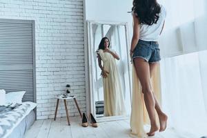 Rückansicht einer attraktiven jungen Frau, die ihr Kleid anprobiert, während sie zu Hause in den Spiegel schaut foto