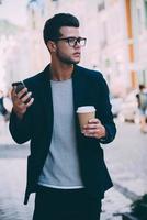 urbaner Lebensstil. hübscher junger Mann in eleganter Freizeitkleidung, der die Straße entlang geht, während er eine Kaffeetasse und ein Smartphone hält foto