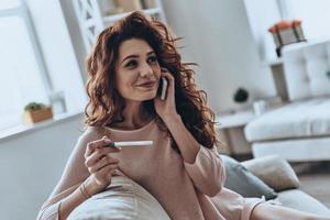 Ratet mal, welche attraktiven jungen Frauen einen Schwangerschaftstest halten und mit ihrem Smartphone sprechen, während sie zu Hause auf dem Sofa sitzen foto