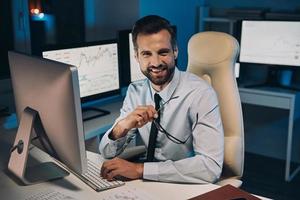 hübscher junger Mann, der seine Brille hält und mit einem Lächeln in die Kamera schaut, während er lange im Büro bleibt foto