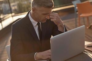 müder reifer geschäftsmann, der die nase massiert, während er draußen auf dem laptop arbeitet foto