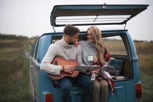 echte Seelenverwandte. hübscher junger mann, der gitarre für seine schöne freundin spielt, während er im kofferraum des blauen minivans im retro-stil sitzt foto