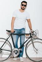 Nur ich und mein Fahrrad. in voller Länge von einem hübschen jungen indischen Mann mit Sonnenbrille, der sich an sein Fahrrad im Retro-Stil lehnt, während er vor weißem Hintergrund steht foto