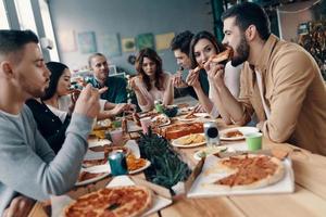 tolles Essen und Gesellschaft. Gruppe junger Leute in Freizeitkleidung, die Pizza essen und lächeln, während sie drinnen eine Dinnerparty haben foto