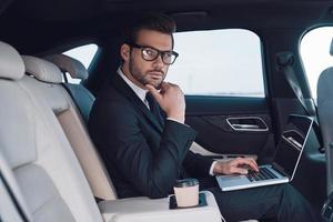 erfolgreicher Profi. nachdenklicher junger Mann im vollen Anzug, der mit Laptop arbeitet und seine Brille anpasst, während er im Auto sitzt foto