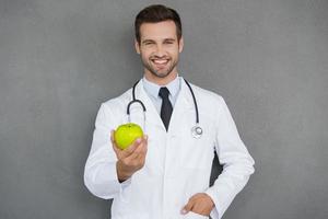 Vitamine sind wichtig für die Gesundheit. Fröhlicher junger Arzt in weißer Uniform, der grünen Apfel ausstreckt und lächelt, während er vor grauem Hintergrund steht foto