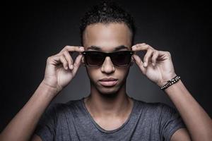 cool und selbstbewusst. Porträt eines jungen afrikanischen Mannes, der seine Sonnenbrille anpasst und in die Kamera schaut, während er vor schwarzem Hintergrund steht foto