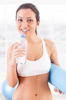 Entspannung nach dem Training. Fröhliche junge Frau in Sportkleidung, die eine Flasche mit Wasser und eine Trainingsmatte hält, während sie im Sportverein steht foto