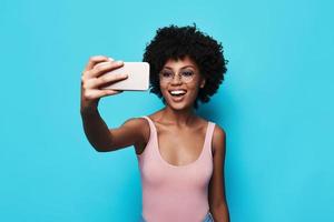 unmöglich, ihrer Schönheit zu widerstehen. Attraktive junge Afrikanerin lächelt und macht Selfie, während sie vor blauem Hintergrund steht foto