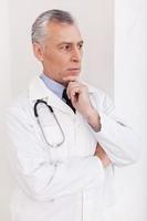 nachdenklicher männlicher Arzt. leitender Arzt mit grauem Haar in Uniform, der wegschaut und die Hand am Kinn hält foto