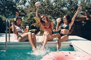 Attraktive junge Frauen im Bikini, die mit erfrischenden Cocktails anstoßen und lächeln, während sie draußen am Pool sitzen foto