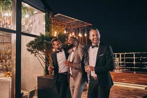 drei glückliche männer in anzügen, die gläser mit whisky halten, während sie zeit auf der party verbringen foto