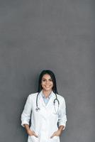 selbstbewusste Ärztin. Selbstbewusste junge Ärztin im weißen Laborkittel, die Hände in den Taschen hält und lächelt, während sie vor grauem Hintergrund steht foto