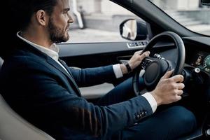 vorsichtiges Fahren. hübscher junger Mann im vollen Anzug, der geradeaus schaut und lächelt, während er ein Auto fährt foto