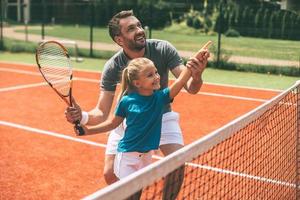 Tennis macht Spaß, wenn der Vater in der Nähe ist. Fröhlicher Vater in Sportkleidung bringt seiner Tochter das Tennisspielen bei, während beide auf dem Tennisplatz stehen foto
