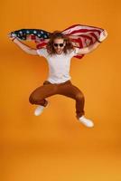 Fröhlicher junger Mann in Freizeitkleidung, der ein Gesicht macht und in die Kamera schaut, während er die amerikanische Flagge vor gelbem Hintergrund hält