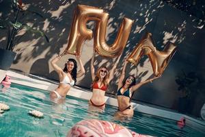 Attraktive junge Frauen in Badebekleidung lächeln und heben Luftballons hoch, während sie draußen im Pool stehen foto