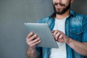 Die Zukunft liegt in der Berührung. Zugeschnittenes Bild eines jungen Mannes, der ein digitales Tablet hält und lächelt, während er vor grauem Hintergrund steht foto